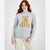 A woman wearing a light gray crewneck sweater with a knitted teddy bear graphic on the chest, paired with beige cargo pants, against a plain light gray background.