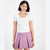 A woman modeling a white short-sleeve scoop-neck t-shirt paired with a lavender pleated mini skirt, set against a plain light gray studio background.