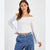A woman modeling a white off-the-shoulder long-sleeve crop top with a tie detail at the neckline, paired with blue jeans, against a plain light gray studio background.