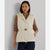 A woman modeling a cream quilted puffer vest with a tan ribbed knit long-sleeve shirt underneath and dark blue jeans, set against a plain light gray background.