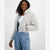 A woman wearing a light gray sherpa fleece cropped puffer jacket over a white top and light blue jeans, standing against a plain light gray studio background.