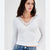 A woman wearing a fitted white long-sleeve ribbed top with a lace V-neckline, paired with blue jeans, against a plain light gray studio background.