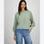 A woman wearing a sage green ribbed knit sweater with balloon sleeves and light blue jeans, standing against a light gray studio background.
