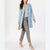 A woman models a light blue mid-length coat over a gray top and gray plaid trousers, holding a small tan clutch against a clean white studio background.