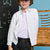A young boy wearing a white long-sleeve button-up shirt over a white t-shirt, dark jeans, and a beige baseball cap, standing in front of a blue wall with a chalkboard and wooden abacus in the background.