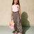 A young girl stands against a textured beige wall wearing a light pink sleeveless top and leopard-print wide-leg pants, holding a pink and beige striped straw bag, and wearing pink sandals.
