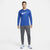 A man modeling a royal blue long-sleeve Nike athletic shirt with a white swoosh logo, paired with gray athletic pants and white running shoes, against a light gray studio background.