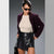 A woman modeling a dark plum corduroy jacket, cream knit top, and black faux-leather wrap mini skirt against a gray studio background.