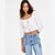A woman wearing a white cropped blouse with lace-up front and eyelet details, paired with light blue jeans, against a plain white studio background.