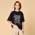 A smiling young boy wearing a black oversized graphic t-shirt with a distressed white 'NYC NEVER STOP ROLLING' skateboard design, paired with beige pants, set against a solid beige studio background.