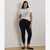 A woman stands against a plain off-white wall wearing a white graphic t-shirt, dark navy blue skinny jeans, and black flats.