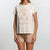 A woman wearing a cream-colored short-sleeve t-shirt with a rose gold graphic of pumpkins and the text 'Pumpkin Spice Brrr' paired with beige shorts, set against a plain light gray background.