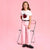 A young girl stands on a pink studio background wearing a white graphic T-shirt with a red ladybug print and the words 'I'm busy', paired with light pink wide-leg pants, a black belt, and black loafers. She is posed next to a clear plastic folding chair.