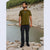 A man stands on rocks by a river wearing an olive green t-shirt with a small blue and white chest graphic, black pants, and black shoes. The background shows a rocky shoreline and green trees.