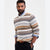 A man wearing a long-sleeve knit sweater with horizontal multicolor geometric stripes in beige, brown, black, and white, layered over a light blue collared shirt and dark trousers.