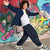 A smiling boy with curly hair poses in front of a colorful graffiti wall, wearing a navy blue zip-up jacket, white graphic t-shirt, navy cargo pants, and green and white sneakers.