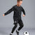 A boy wearing a black long-sleeve athletic top and matching black shorts with white piping details, posed dynamically while kicking a soccer ball on a solid gray studio background.