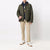 A model wearing an olive green quilted jacket with brown leather shoulder details, layered over a navy hoodie, beige trousers, and white sneakers, standing against a plain white studio background.
