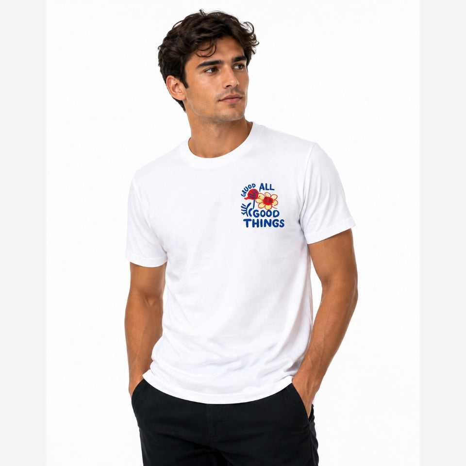 A man wearing a white short-sleeve crewneck t-shirt with a colorful graphic on the left chest that reads 'ALL GOOD THINGS' with a small rainbow design, paired with black pants, against a plain white background.