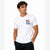A man wearing a white short-sleeve crewneck t-shirt with a colorful graphic on the left chest that reads 'ALL GOOD THINGS' with a small rainbow design, paired with black pants, against a plain white background.