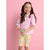 A young girl with brown pigtails stands against a solid pink background, wearing a light pink ruffled button-up shirt and cream-colored capri leggings with a yellow lemon print. She is holding a small teal ball.