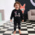 A toddler stands barefoot on a black-and-white checkered floor wearing a black two-piece pajama set with a monster face graphic on the sweatshirt and matching pants.