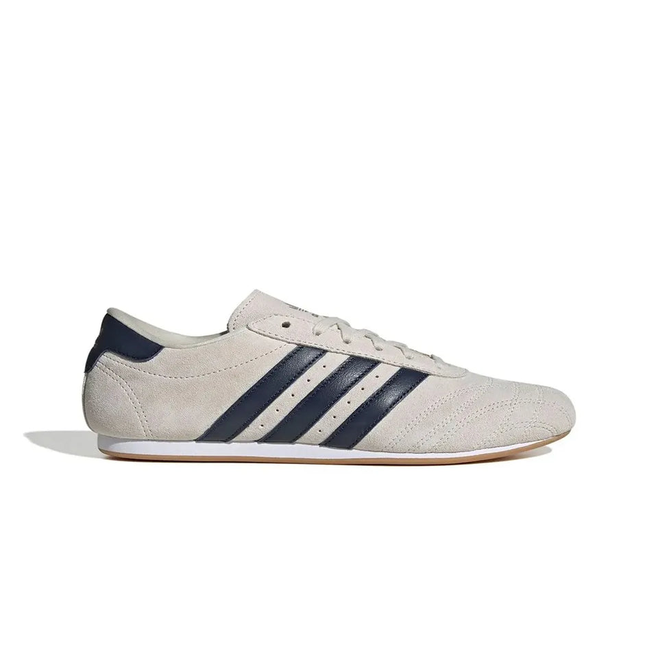 A light beige suede sneaker with navy blue three-stripes on the side, white laces, and a white midsole with a gum outsole, shown in profile against a white background.