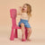 A young child with blonde hair sits happily on a small pink plastic chair against a solid beige background. The child is wearing a beige t-shirt and a blue denim skirt.