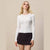 A blonde woman modeling a fitted white ribbed long-sleeve top with a front button closure, paired with a dark skirt, against a plain beige studio background.