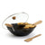 A black wok with a clear glass lid, wooden handles, and a metal rack holding two pieces of golden fried food, shown on a white background with a wooden spatula nearby.
