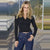 A woman modeling a fitted black long-sleeve V-neck crop top with dark blue jeans, standing on a boardwalk with a blurred outdoor background.