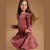 A young girl with long light brown hair is wearing a dusty rose velvet dress with small white star patterns, posing against a solid warm brown studio background.