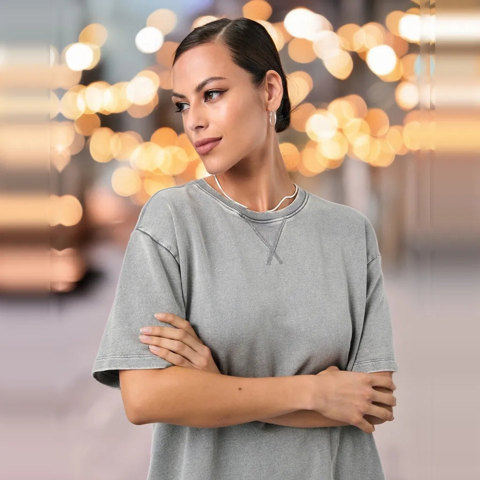 A woman wearing a light gray oversized crewneck sweatshirt, standing with her arms crossed against a blurred background of warm bokeh lights.