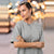 A woman wearing a light gray oversized crewneck sweatshirt, standing with her arms crossed against a blurred background of warm bokeh lights.