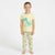 A young girl stands barefoot against a plain white studio background wearing a pale yellow pajama set with a large blue dinosaur graphic on the shirt and multicolored dinosaur prints on the pants.
