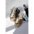 A pair of metallic gold slide sandals with wide straps and square toes, styled on top of a magazine page against a light gray background with soft shadows.