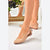 A pair of women's pointed-toe flat shoes in a light nude beige color, shown on a model's feet. The shoes feature a low block heel and a delicate ankle strap with a bow detail.