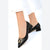 A pair of black pointed-toe pumps with a gold-tone V-shaped metal accent on the toe box, shown on a model wearing white pants against a white background.