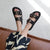 A person wearing black double-strap slide sandals with large gold-tone buckles and gold eyelet details, seated on a brown leather couch. The person is also wearing a navy and cream patterned skirt.