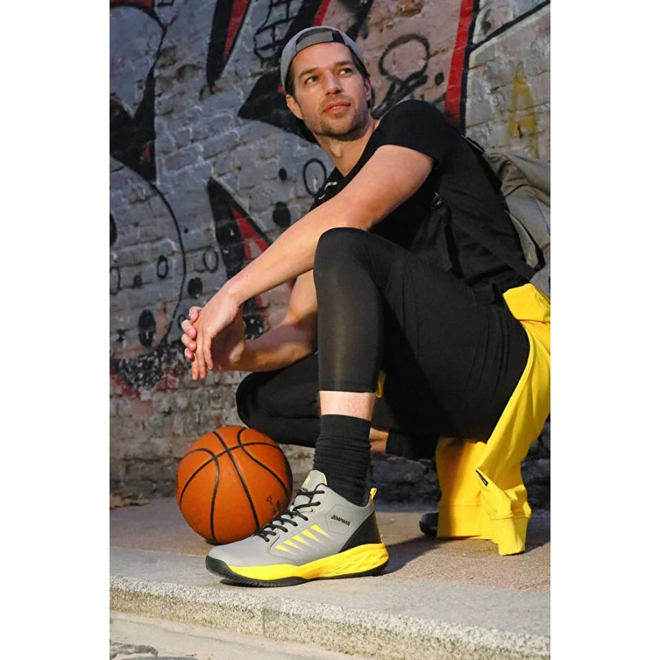 A man in athletic wear crouches on a concrete ledge in front of a graffiti-covered brick wall, with a basketball at his side. He is wearing a black t-shirt, black leggings, a gray and yellow running shoe, and a yellow jacket.