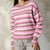 A person wearing a pink, white, and mauve striped knit sweater with gray pants, standing against a textured gray wall.