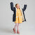 A young girl with blonde hair is smiling and posing with her arms raised, wearing a navy blue puffer jacket with a cream sherpa lining over a yellow polka-dot dress, pink tights, and pink sneakers, set against a plain light gray studio background.