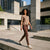 A woman walking outdoors in a modern concrete urban setting, wearing a taupe short-sleeve knit top and matching taupe leggings with white sneakers.