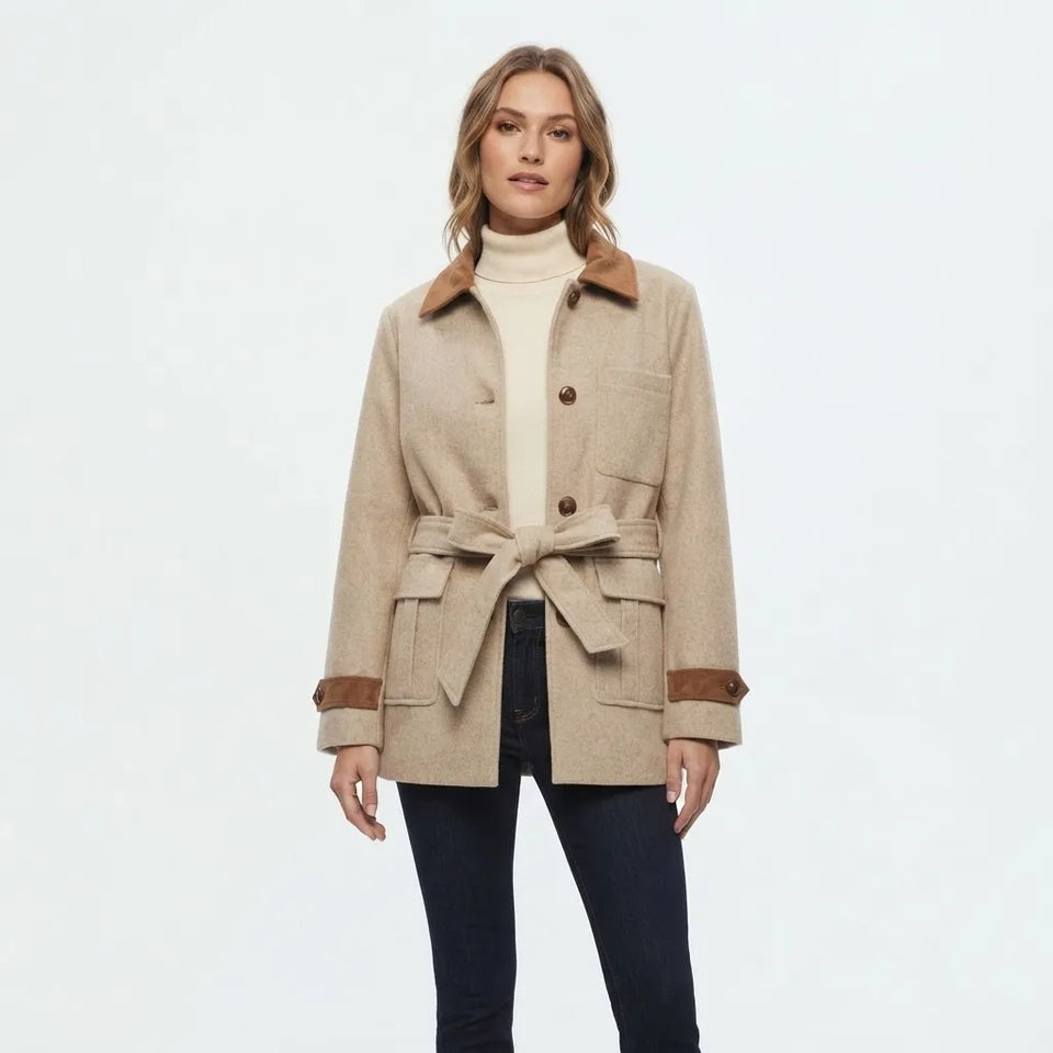 A woman modeling a beige wool-blend coat with brown suede-like collar, cuffs, and pocket trim, worn over a cream turtleneck and dark jeans, against a plain light gray studio background.
