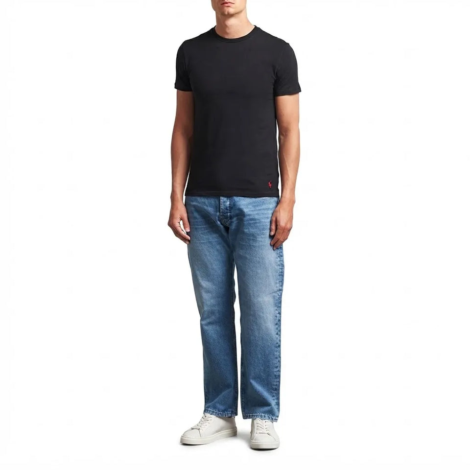 A man wearing a black short-sleeve crewneck t-shirt, blue jeans, and white sneakers, standing against a plain white background.