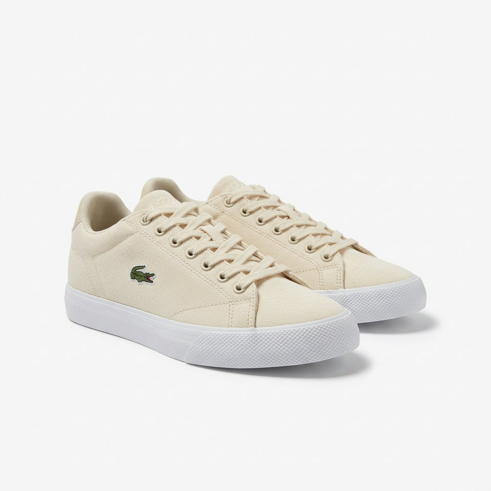 A pair of Lacoste low-top sneakers in a light cream canvas, featuring the signature green crocodile logo on the side and white rubber soles, set against a plain light gray background.