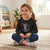 A young girl with pigtails sits cross-legged on a woven rug in a bright room, wearing a black long-sleeve ruffle-sleeve shirt with a gray cat and pink bow graphic, blue jeans, and gray sneakers.