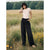 A woman stands in a grassy field with wildflowers and trees in the background, wearing a fitted light beige short-sleeve top and loose black wide-leg jeans.