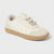 A low-top sneaker in off-white suede and leather, featuring decorative silver-tone rhinestone and pearl laces, a cream collar, and a tan rubber sole, shown on a light gray background.