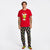 A man modeling a red short-sleeve t-shirt and dark navy patterned pajama pants with a Grinch-themed holiday print, standing against a plain white studio background.